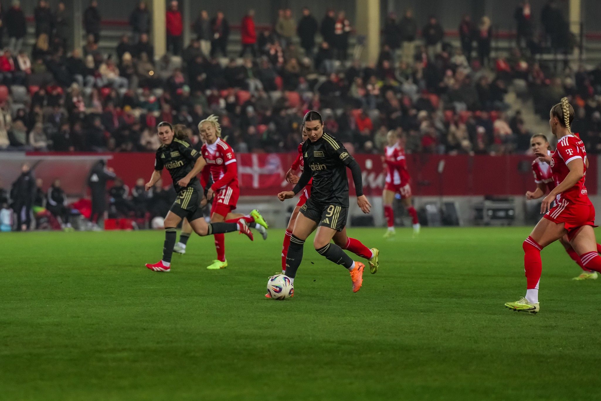 Spielszene eines Fußballspiels mit mehreren Spielern auf dem Feld, im Hintergrund Zuschauer auf Tribünen.