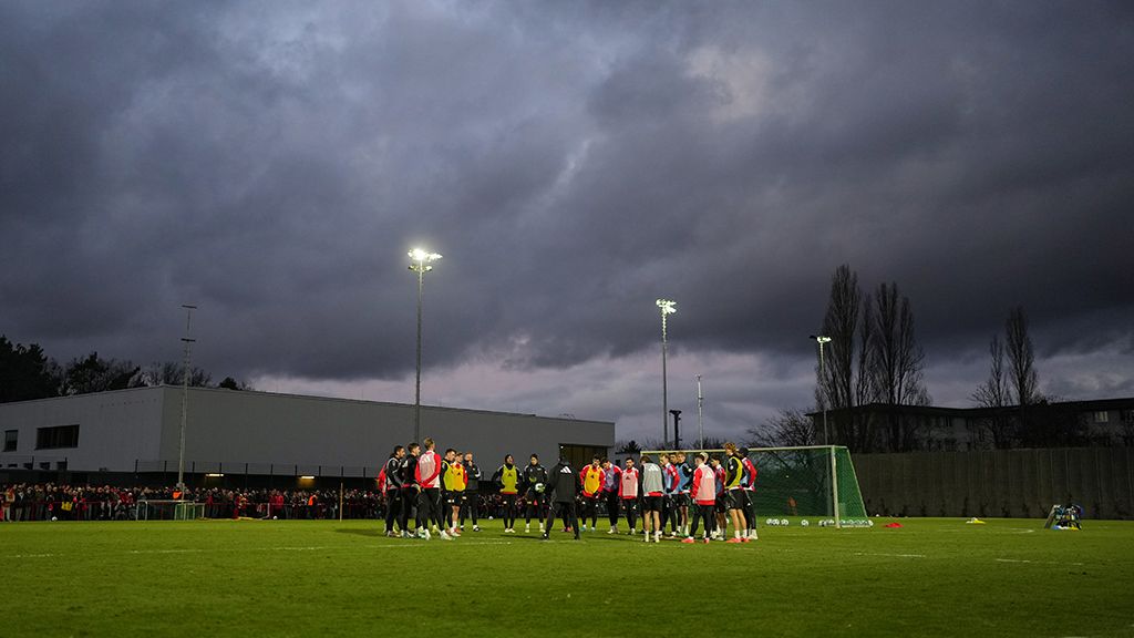 Fußballer in Trainingsanzügen versammeln sich auf einem grünen Platz unter bewölktem Himmel mit schwachem Licht.