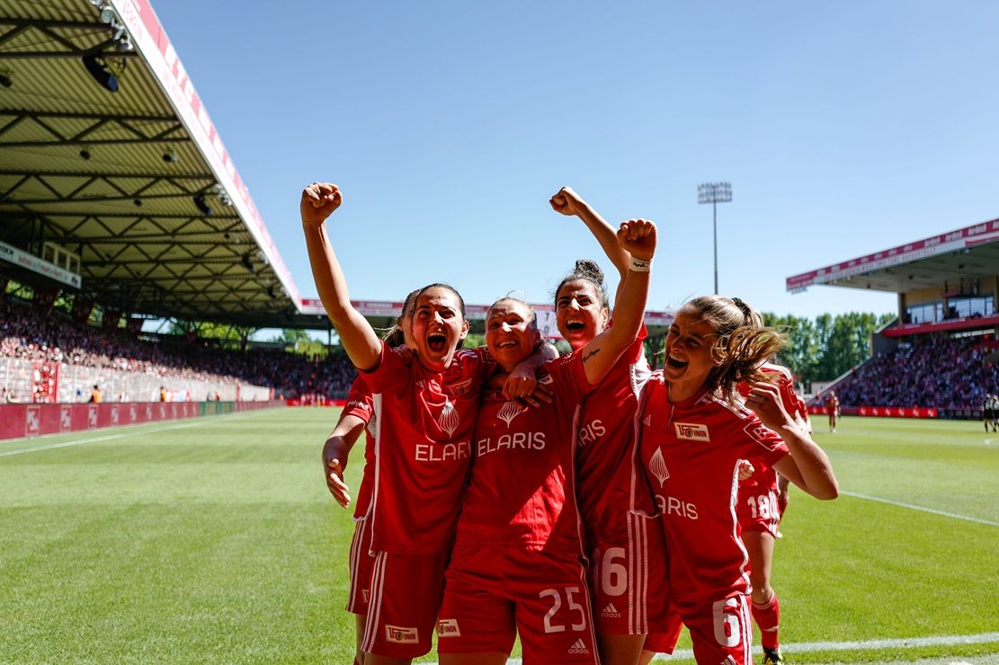 Vier jubelnde Fußballspielerinnen in roten Trikots feiern einen Sieg auf dem Spielfeld, umgeben von einem begeisterten Publikum.