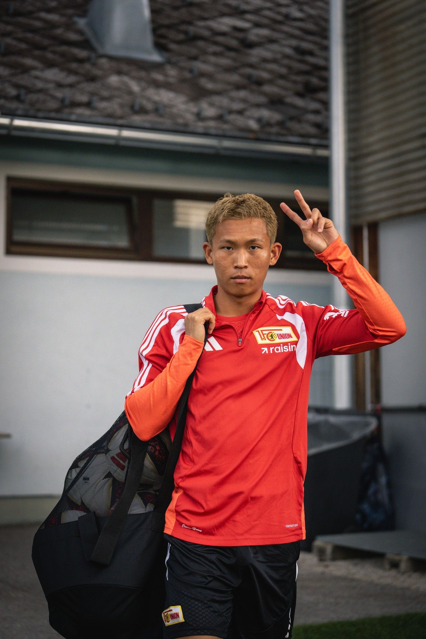 Junger Mann mit kurzem blondem Haar in rotem Sporttrikot, der eine Tasche trägt und mit zwei hochgestellten Fingern posiert.