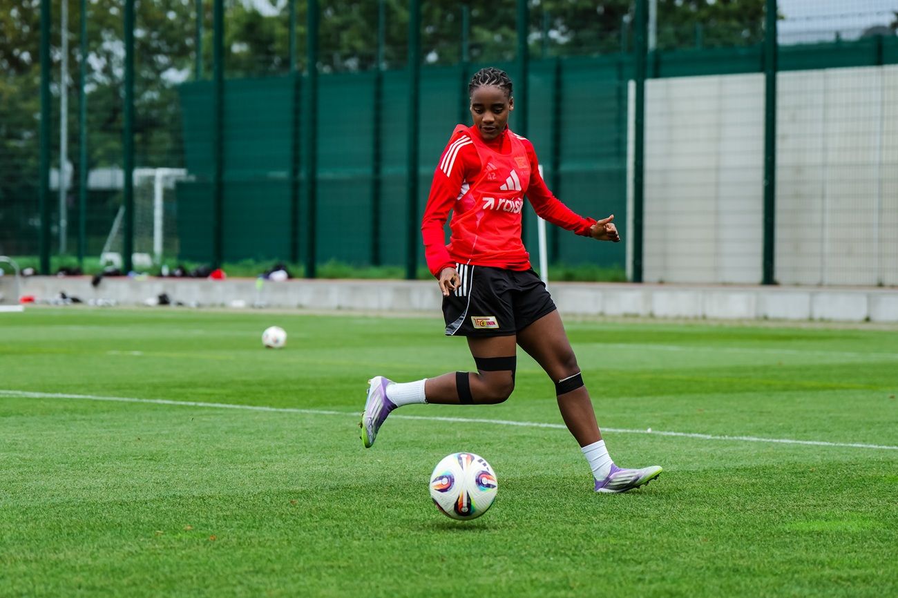 Eine Fußballspielerin in rotem Trikot tritt einen Ball auf einem Trainingsplatz mit Kunstrasen.