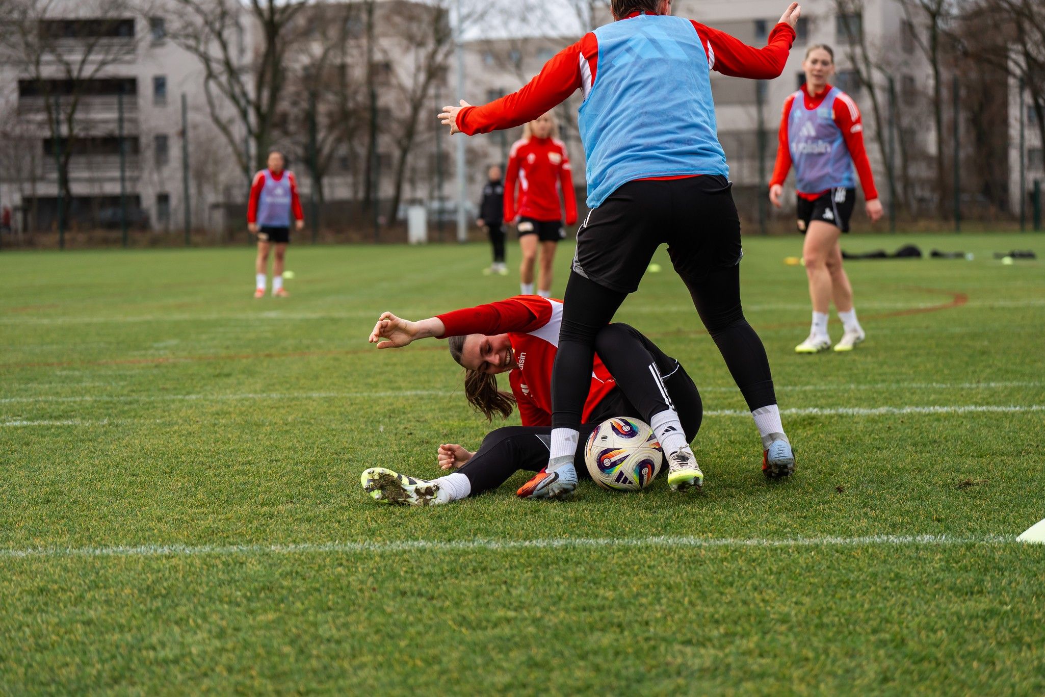 Zwei Frauen im roten Trikot kämpfen um den Ball auf dem Fußballfeld, während andere Spielerinnen im Hintergrund trainieren.