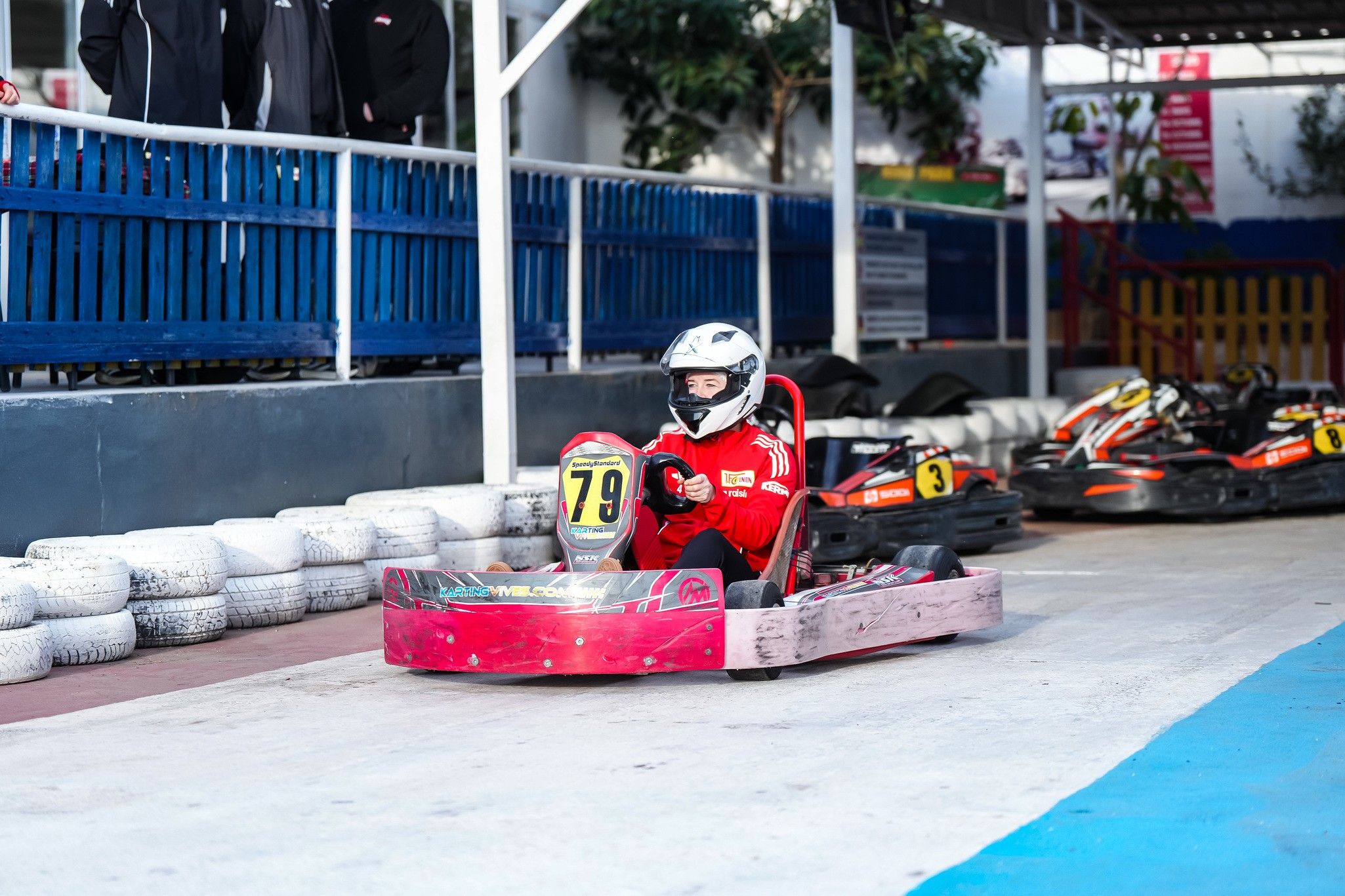 Ein Kind in roter Rennkleidung fährt mit einem Go-Kart auf einer Indoor-Rennstrecke. Reifen und andere Karts sind im Hintergrund sichtbar.