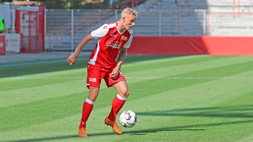 A young soccer player in a red jersey dribbles across the green field during a game.