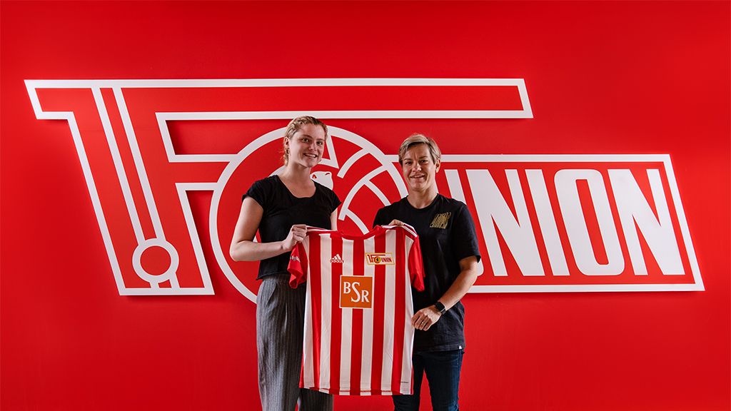 Two people hold a red and white striped jersey in front of a red wall with the logo of 1. FC Union Berlin.