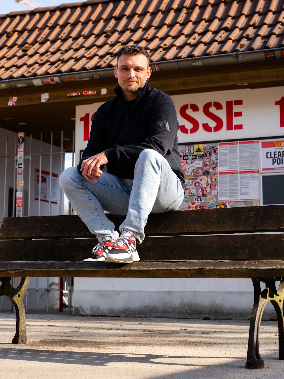 Ein Mann sitzt auf einer Parkbank vor einem Gebäude mit Aufklebern und weißen Plakaten. Er trägt einen schwarzen Hoodie und Jeans.