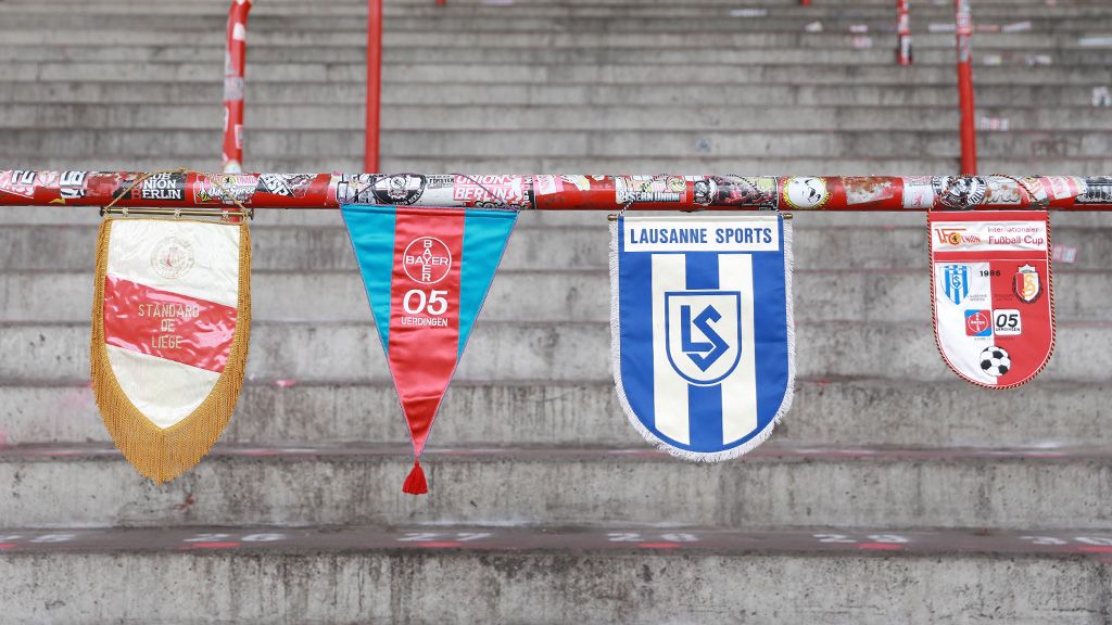 Bunte Fußball-Banner hängen an einem Geländer im Stadion, untermalt von abgenutzten Steinstufen.