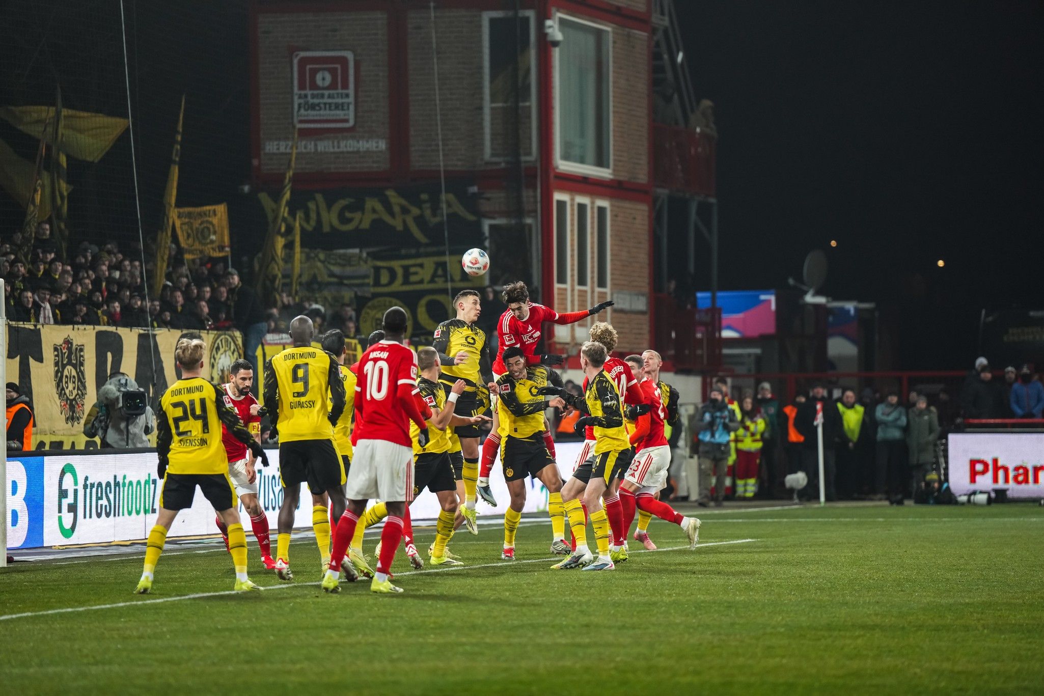 Spielszene mit Spielern in gelben und roten Trikots, die um den Ball kämpfen, während Fans im Hintergrund jubeln.
