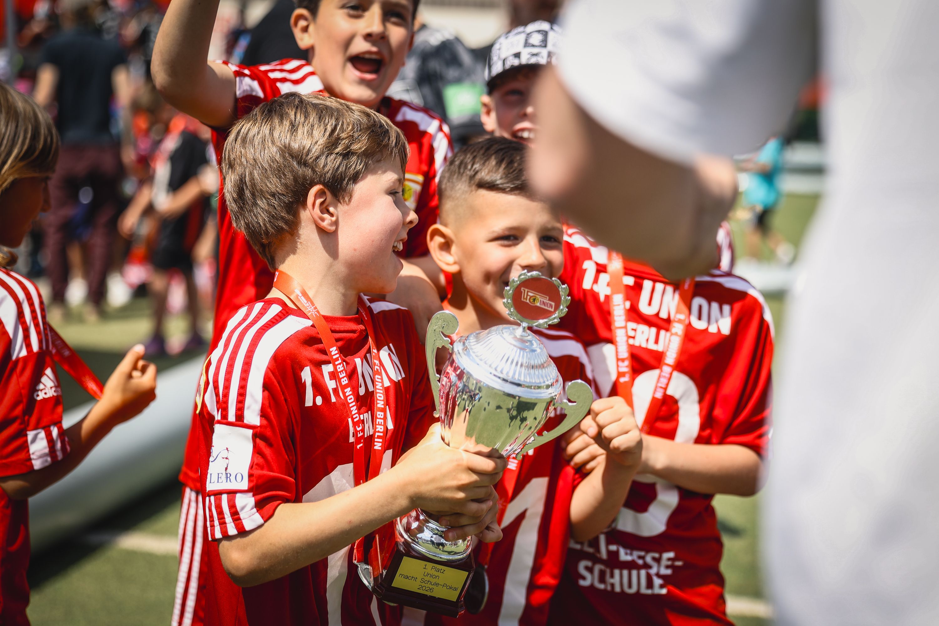 Gruppe von jubelnden Kindern in roten Fußballtrikots, die einen Pokal halten und sich freuen.