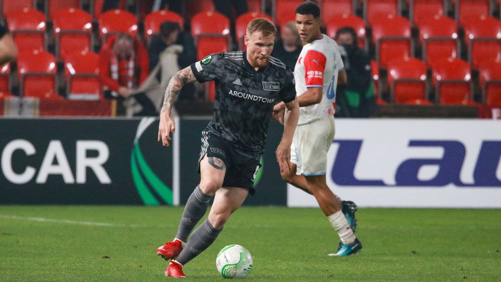 Ein Fußballspieler in einem grauen Trikot dribbelt den Ball während eines Spiels auf einem nassen Platz, im Hintergrund ein anderer Spieler.
