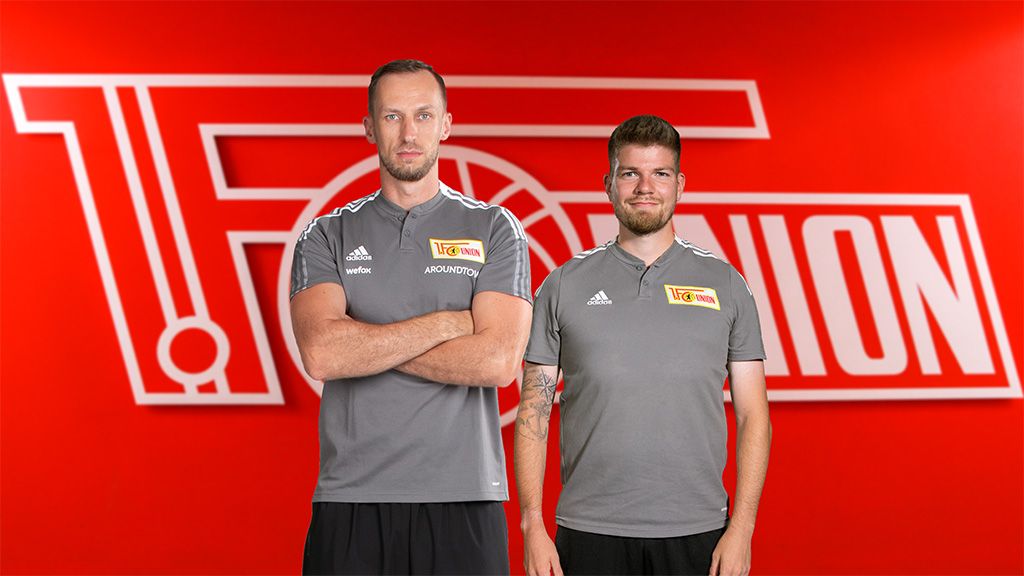 Zwei Männer in grauen Sportshirts stehen vor einem roten Hintergrund mit dem Logo von Union Berlin.