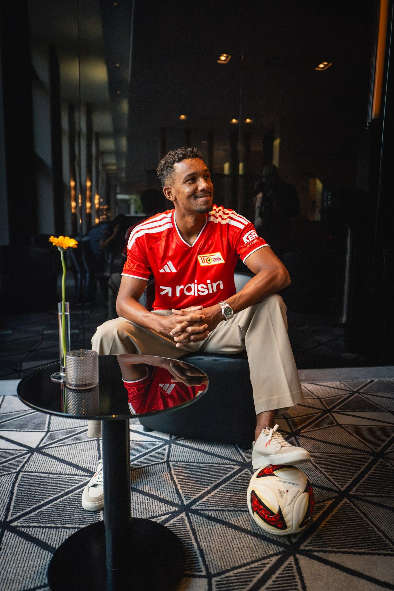 Ein sportlicher Mann in einem roten Fußballtrikot sitzt in einem modernen Raum neben einem Tisch mit einer Blume und einem Fußball.