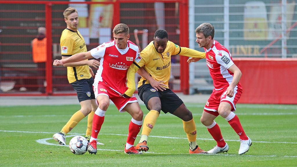Zwei Fußballspieler in rot kämpfen mit einem Spieler in schwarz um den Ball. Sie sind auf einem Fußballfeld.