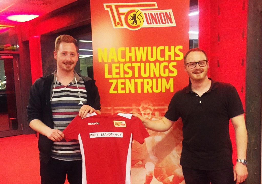 Zwei Männer halten gemeinsam ein Rot-Weiß-Trikot der Fußballmannschaft Union Berlin vor einem Poster des Nachwuchsleistungszentrums.