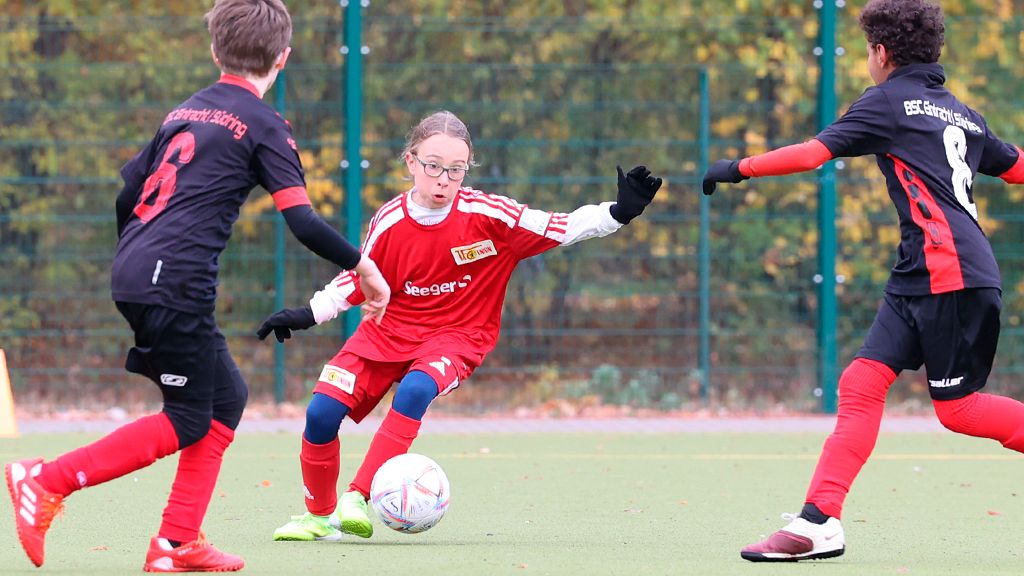 Ein Mädchen im roten Trikot spielt Fußball und dribbelt den Ball zwischen zwei Spielern hindurch.