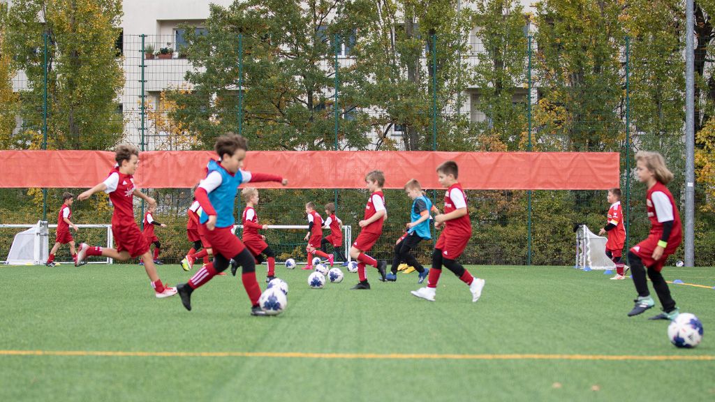 Kinder in roten und blauen Trikots trainieren auf einem Fußballplatz mit mehreren Bällen und einem grünen Kunstrasen.