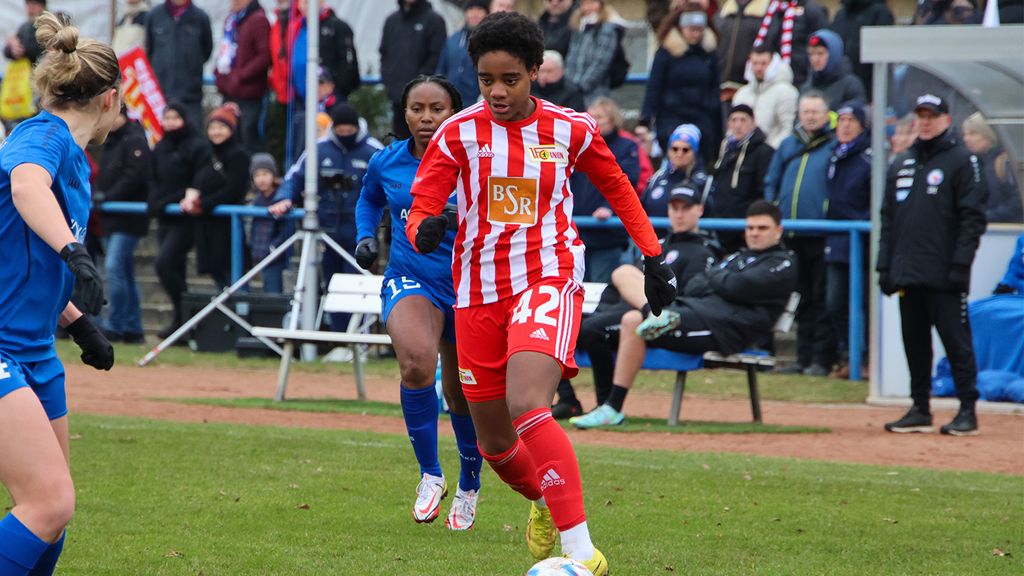A female soccer player in a red and white uniform is dribbling the ball. Spectators in the background.