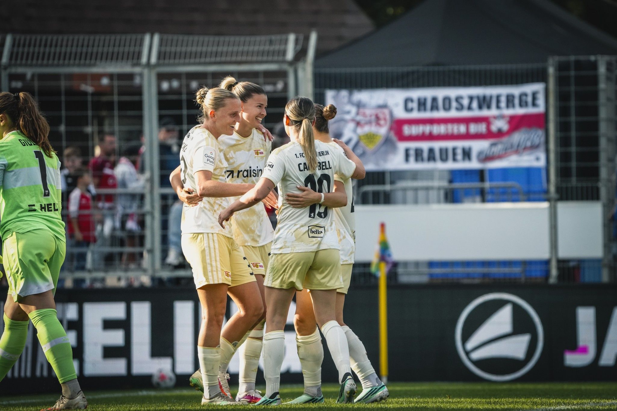 Spielerinnen einer Fußballmannschaft feiern einen Torjubel auf dem Spielfeld, im Hintergrund ein Banner der Fans.