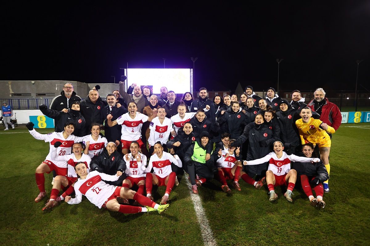 Eine feiernde Gruppe von Frauenfußballspielerinnen und -trainern in roten und weißen Trikots auf einem Sportplatz.