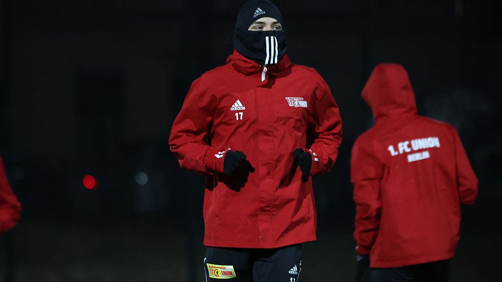 Fußballspieler in rotem Trainingsanzug mit Kapuze und Maske joggt auf dem Platz bei Dunkelheit.