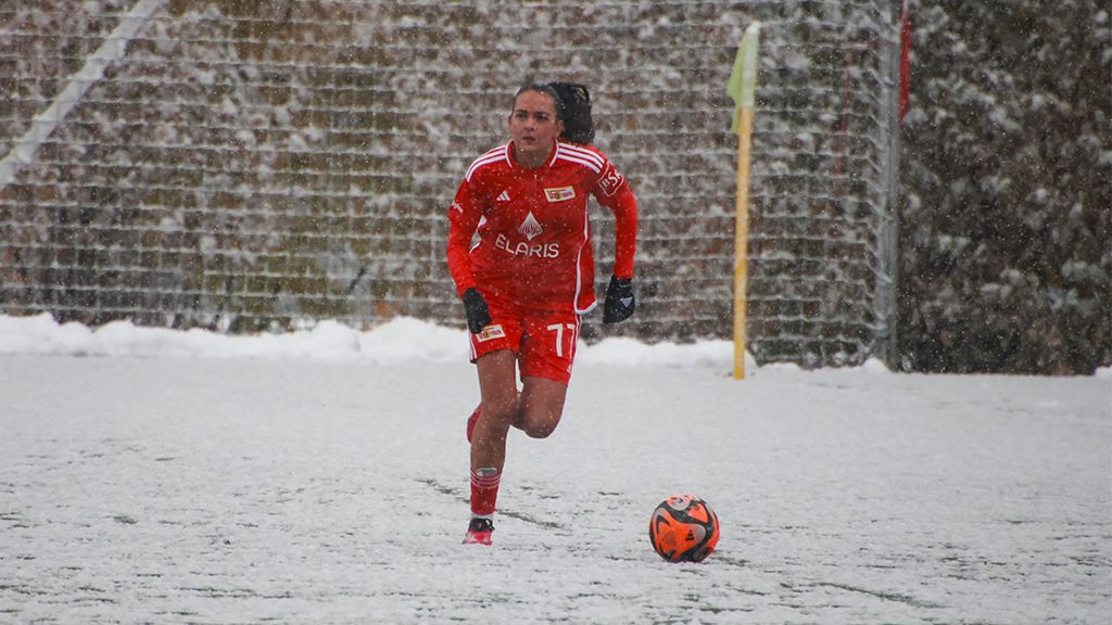 Eine Fußballspielerin in rotem Trikot läuft im Schnee auf einem Spielfeld, während Schneeflocken fallen.