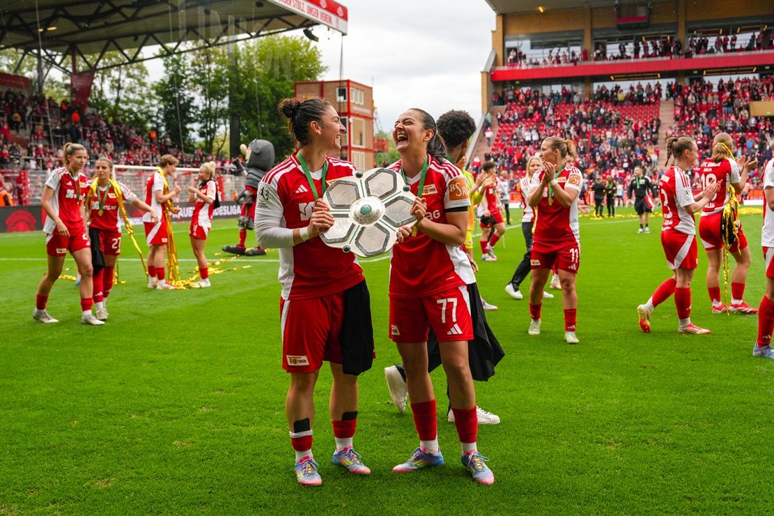 Zwei Spielerinnen in roten Trikots halten stolz einen Pokal auf einem Fußballfeld, umgeben von jubelnden Teamkolleginnen.