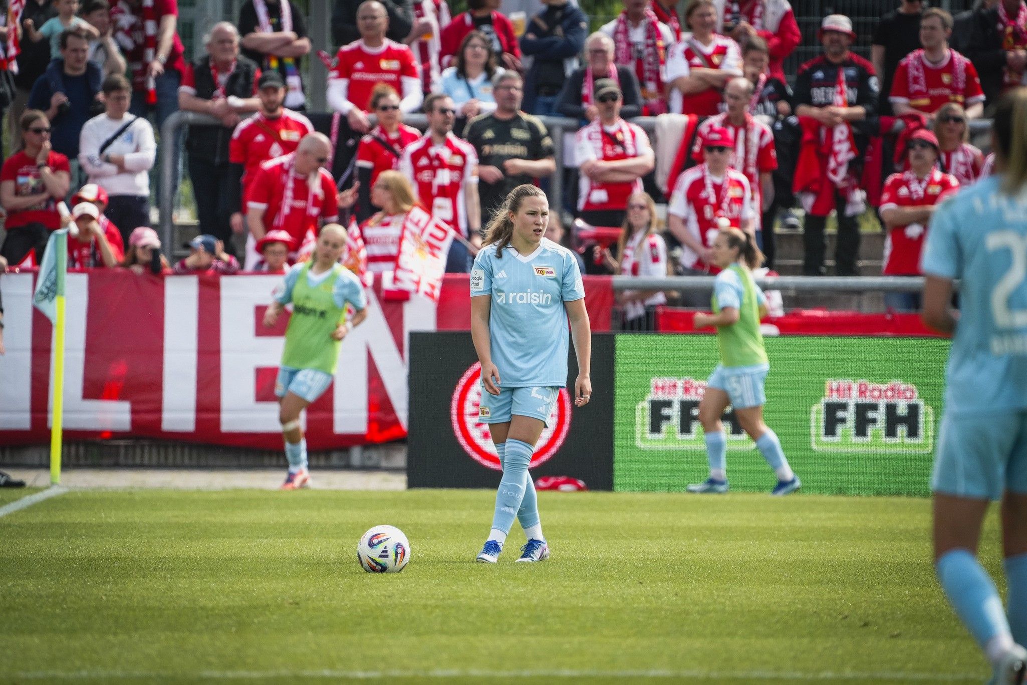 Eine Fußballspielerin in blauer Uniform steht auf dem Spielfeld, während Fans mit roten Trikots im Hintergrund jubeln.