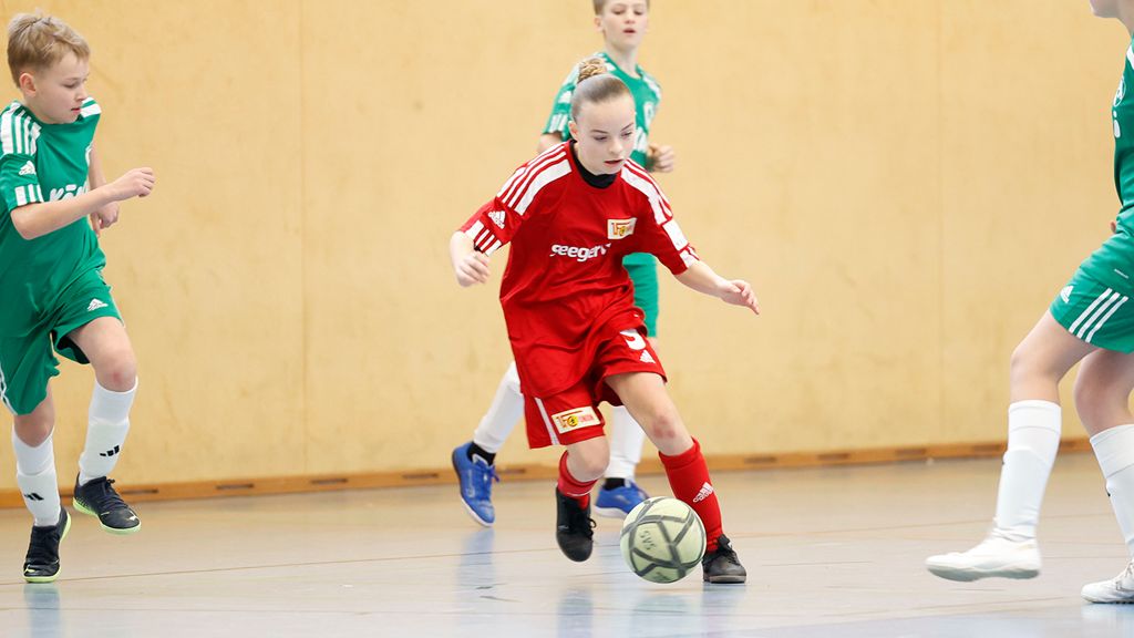 Ein Mädchen in einem roten Fußballtrikot dribbelt den Ball, während zwei Spieler in grünen Trikots auf dem Feld stehen.