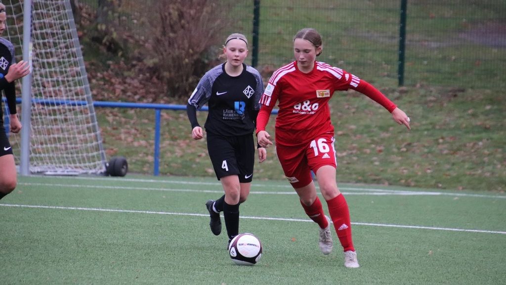 Zwei junge Fußballspielerinnen auf dem Feld, eine in rotem Trikot, die andere in schwarzem Trikot, verfolgen den Ball.