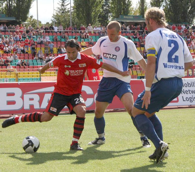 Ein Fußballspiel auf einem Rasenplatz, in dem ein Spieler im roten Trikot versucht, den Ball zu dribbeln, während zwei Gegner ihn umgeben.