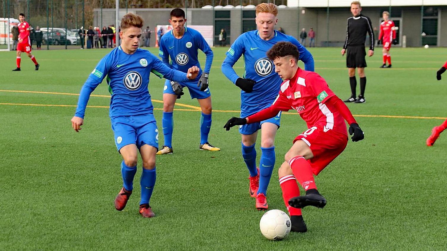 Zwei Spieler in blauen Trikots versuchen, einen Ball von einem Spieler in rotem Trikot zu erobern. Fußballspiel auf einem Platz.