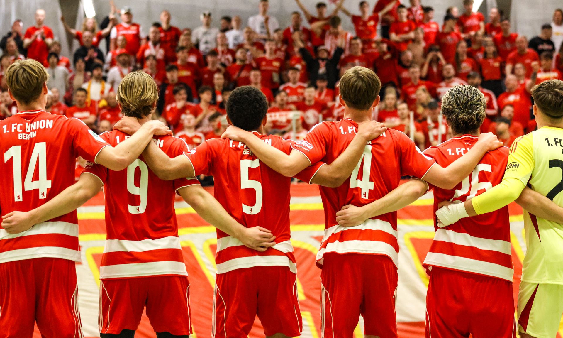 Sechs Fußballspieler in roten Trikots stehen nebeneinander und schauen in Richtung einer jubelnden Menge im Hintergrund.