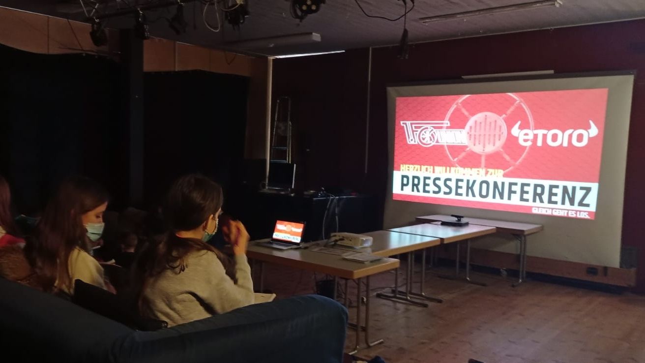 Personen sitzen in einem Raum vor einer Leinwand, auf der das Wort "PRESSEKONFERENZ" und ein Logo angezeigt werden.