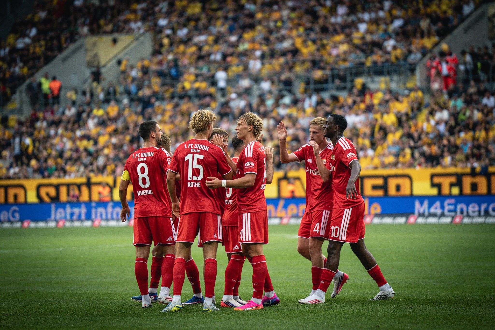 Spieler von FC Köln feiern ein Tor auf dem Spielfeld, umgeben von jubelnden Fans im Hintergrund.