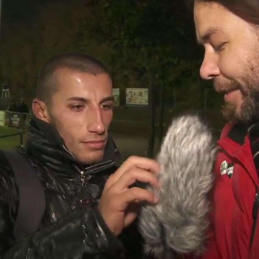Ein Mann mit kurzes Haar in einer schwarzen Jacke berührt ein Mikrofon, während ein anderer Mann in roter Jacke heimlich spricht.
