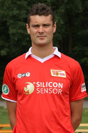 Ein Mann in einem roten Fußballtrikot mit dem Logo "Silicon Sensor" steht vor einer grünen Kulisse.