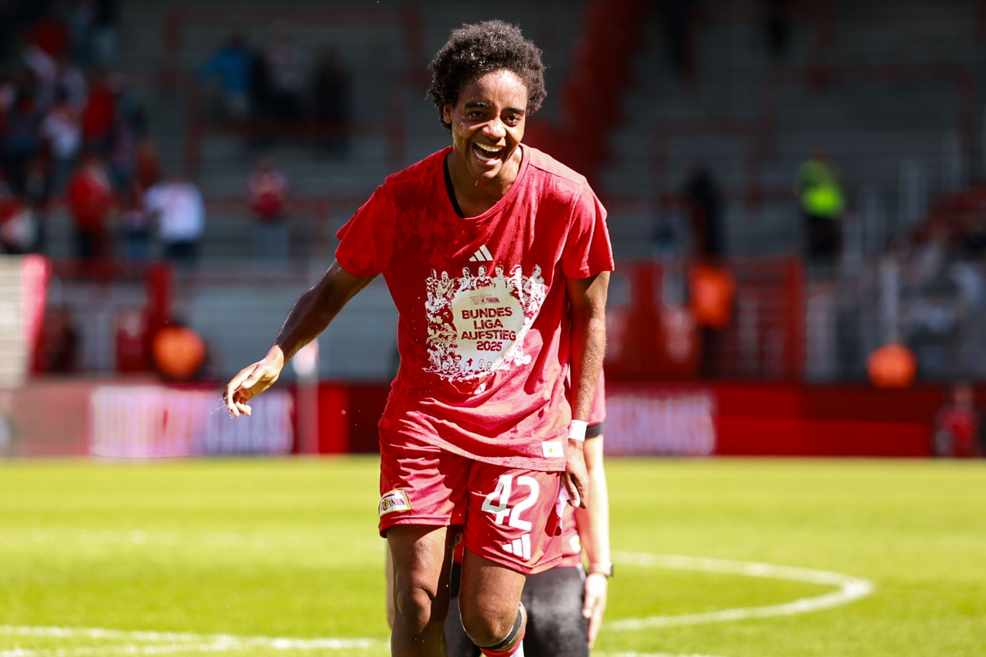 Ein junger Fußballspieler in rotem Trikot mit der Nummer 42 lächelt und feiert auf dem Spielfeld nach einem Spiel.