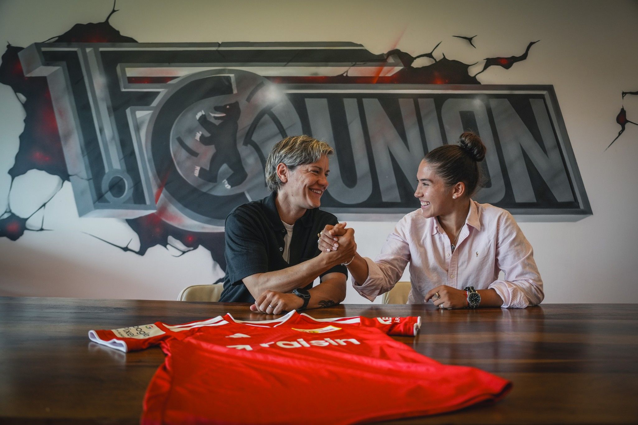 Zwei Personen schütteln sich die Hände vor einem graffitiartigen Hintergrund, mit einem roten Fußballtrikot auf dem Tisch.