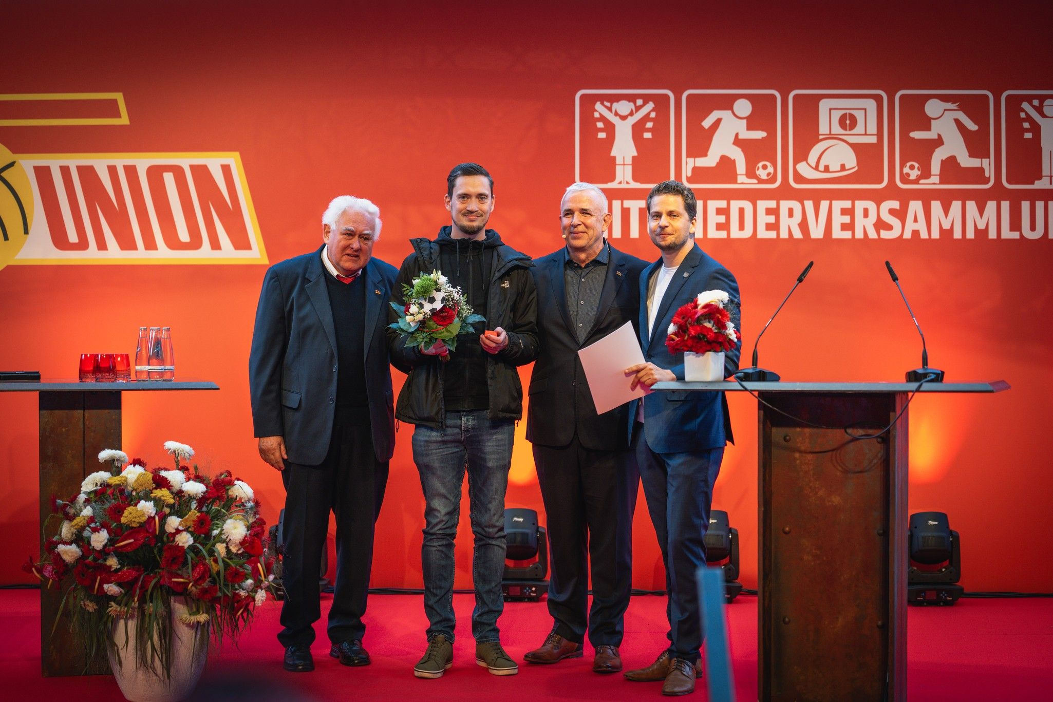 Vier Personen stehen auf einer Bühne, halten Blumen und Dokumente, umgeben von roten Pflanzen und einer dekorativen Hintergrundwand.
