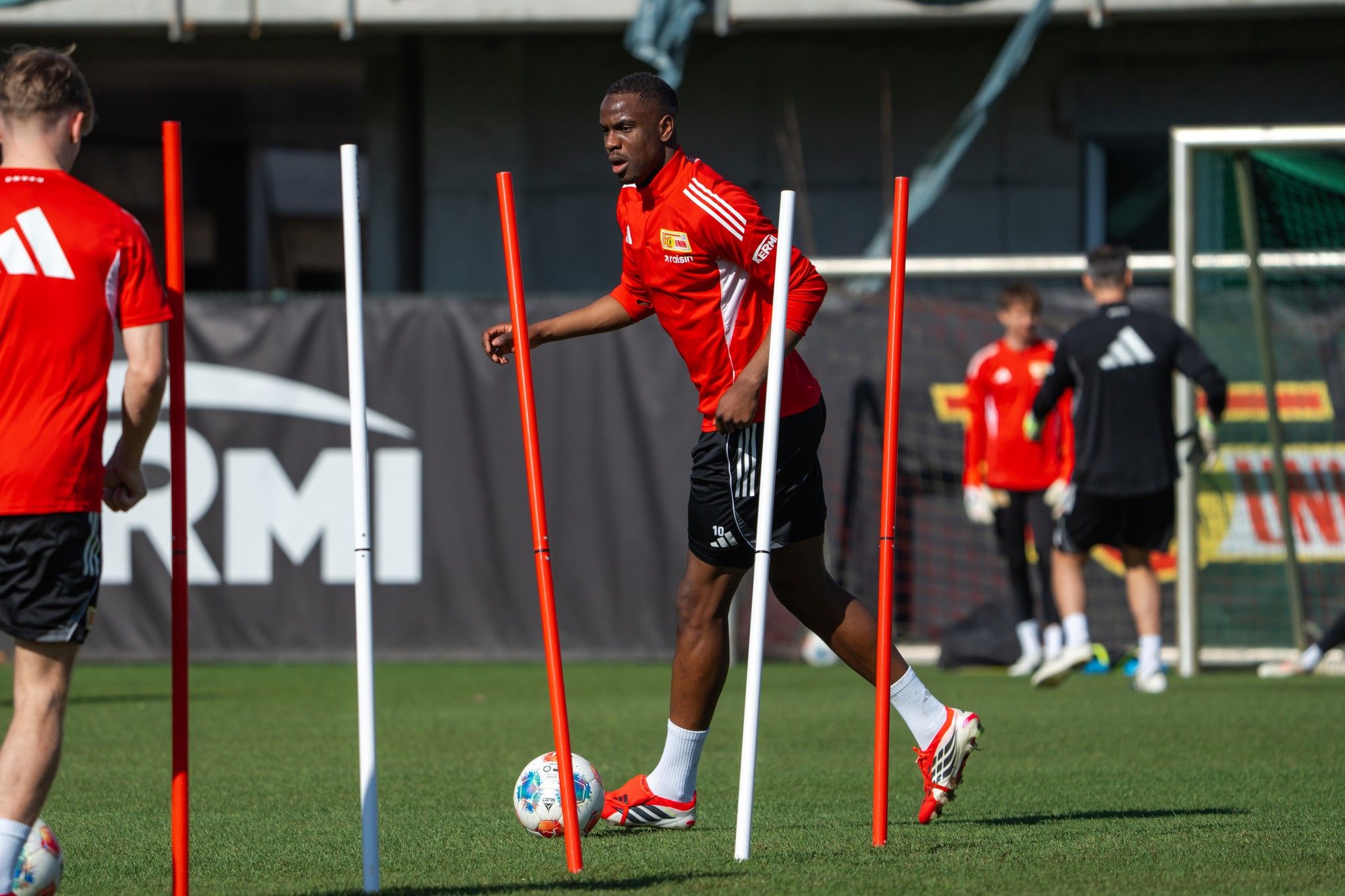 Ein Fußballspieler trainiert auf dem Platz und umgeht hohle Stangen, während andere Spieler im Hintergrund üben.