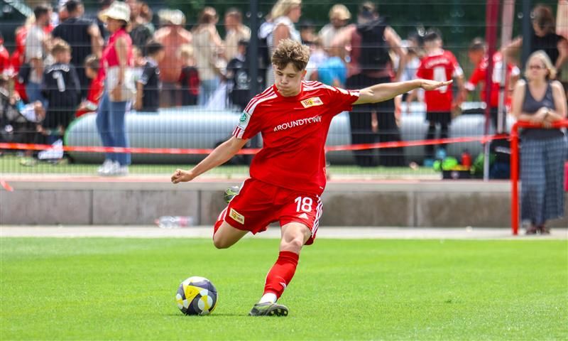 Ein junger Fußballspieler in rotem Trikot schießt den Ball auf einem Rasenplatz während eines Spiels. Zuschauer im Hintergrund.