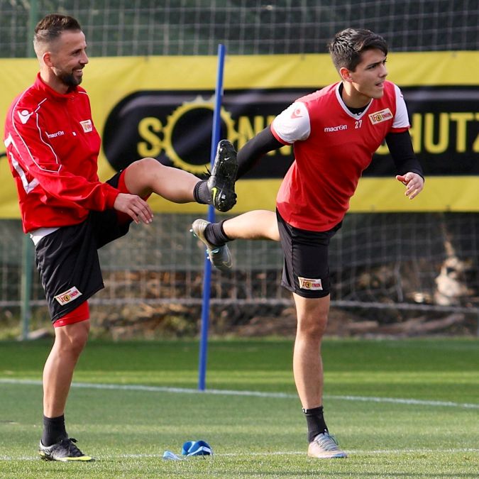 Zwei Fußballspieler in Trainingskleidung, einer hebt sein Bein, während der andere ihn an der Wade unterstützt.