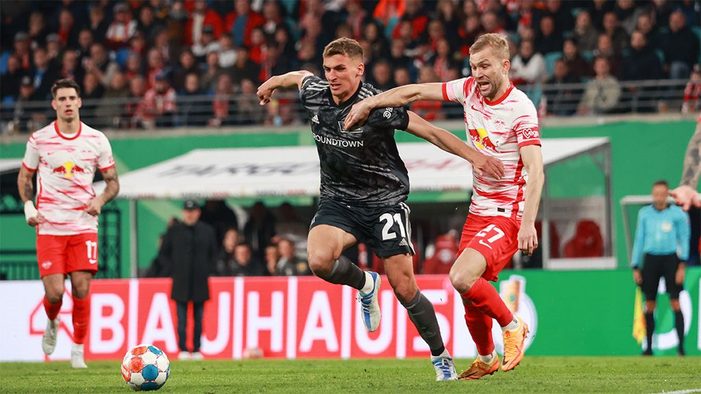 Zwei Fußballspieler kämpfen um den Ball während eines Spiels, mit Zuschauern im Hintergrund.