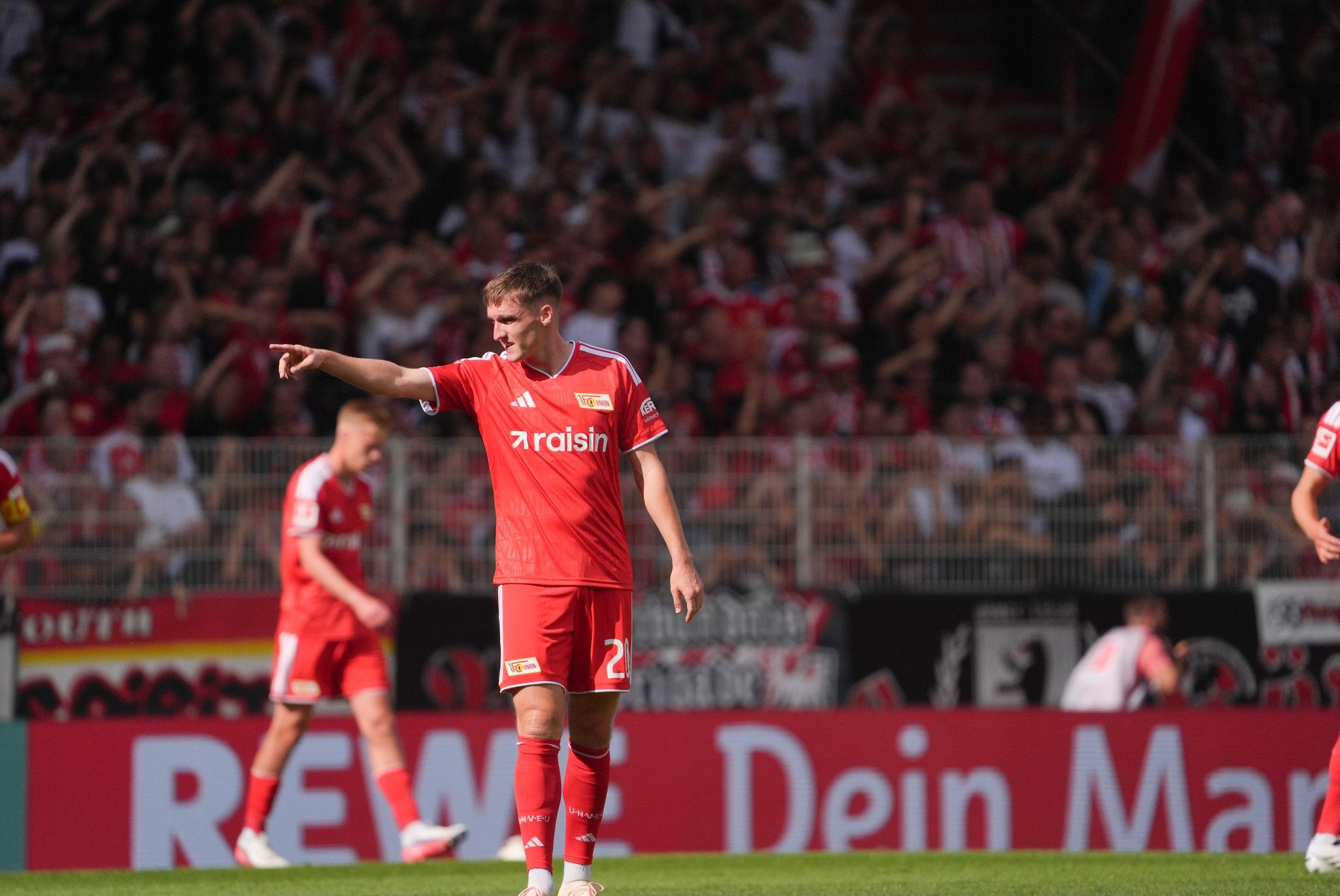 Spieler des 1. FC Union Berlin in rotem Trikot zeigt auf das Spielfeld, im Hintergrund jubelnde Fans.