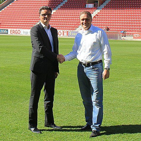 Zwei Männer schütteln sich die Hände auf einem Fußballplatz vor der Tribüne.