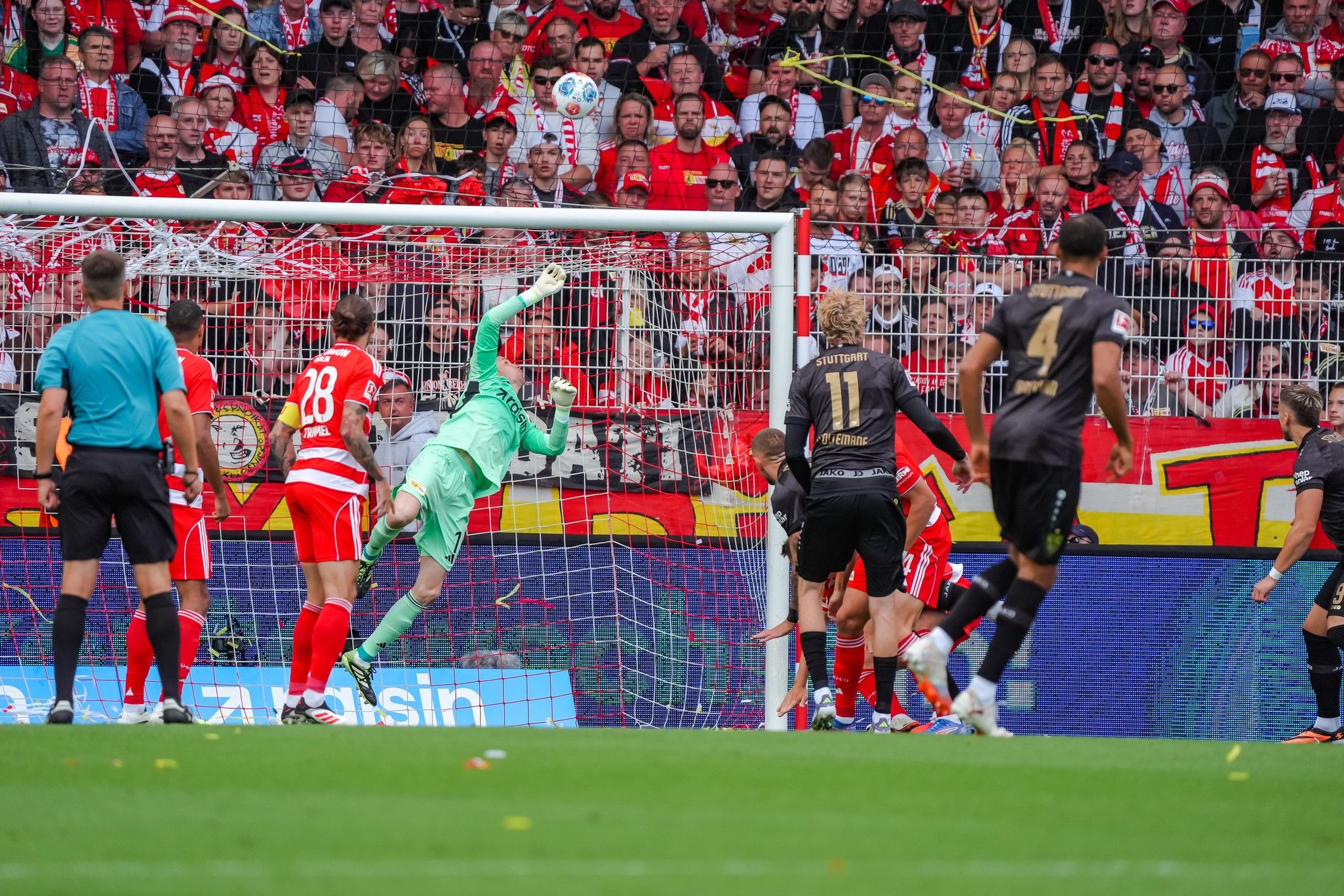 Torwart springt, um einen Ball in einem Fußballspiel abzuwehren, während Fans im Hintergrund jubeln.