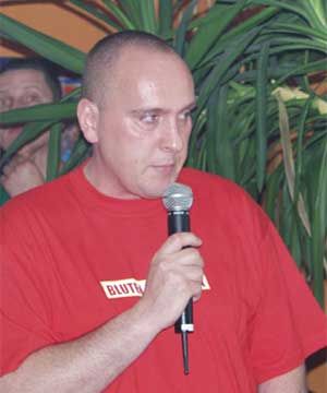 Mann in rotem T-Shirt hält ein Mikrofon, steht vor Pflanzen im Hintergrund.