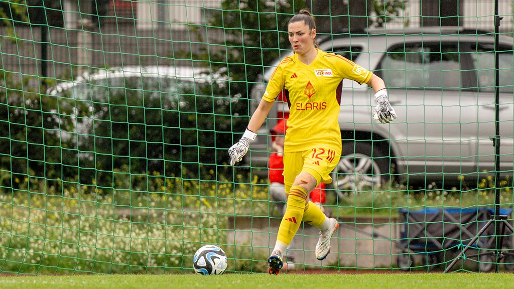 Torhüterin in gelb im Moment des Spiels, steht im Tor und bereitet sich auf einen Schuss vor. Im Hintergrund ist ein Auto sichtbar.