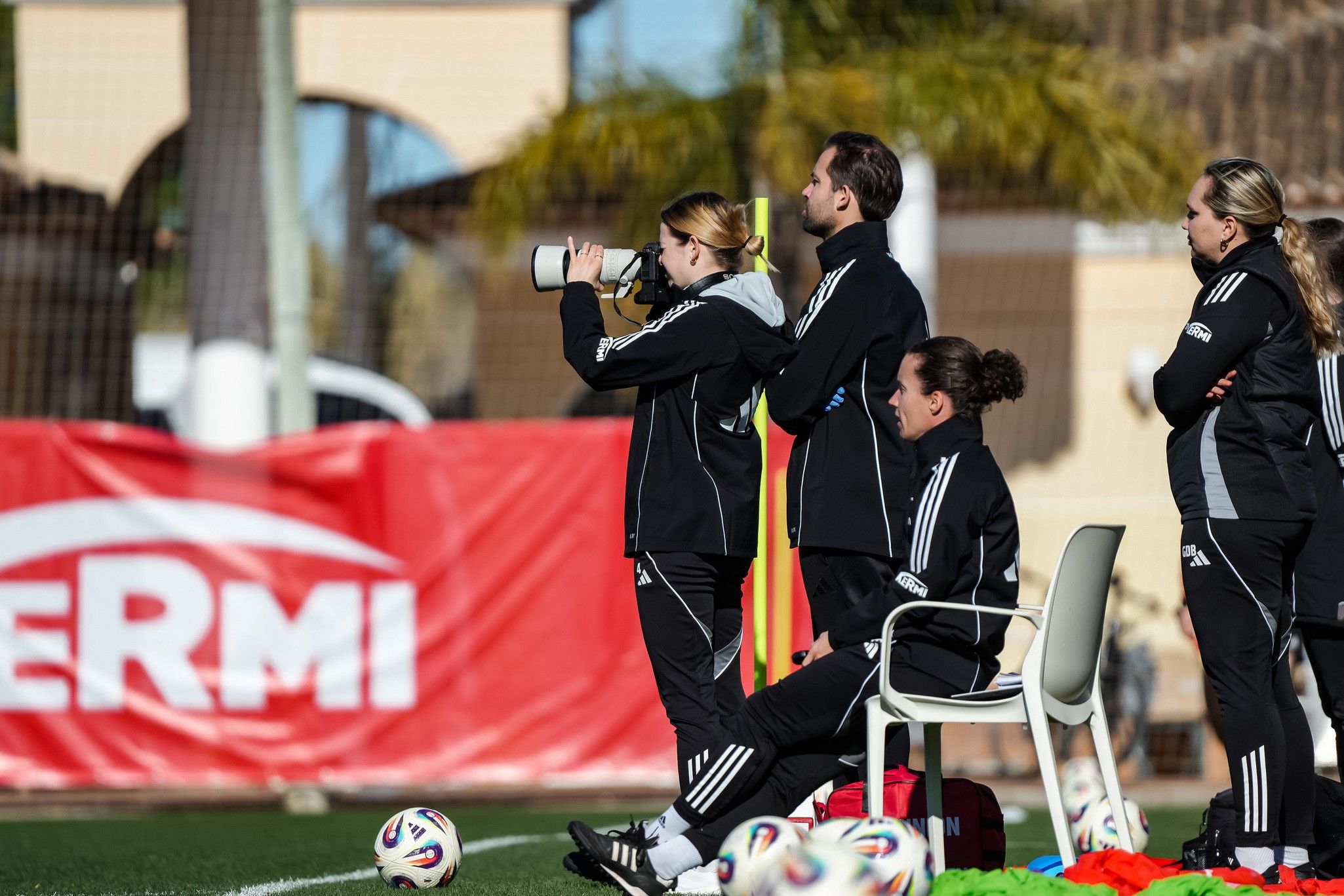 Trainerteam beobachtet ein Fußballspiel, während Spieler umliegende Bälle auf dem Boden liegen.