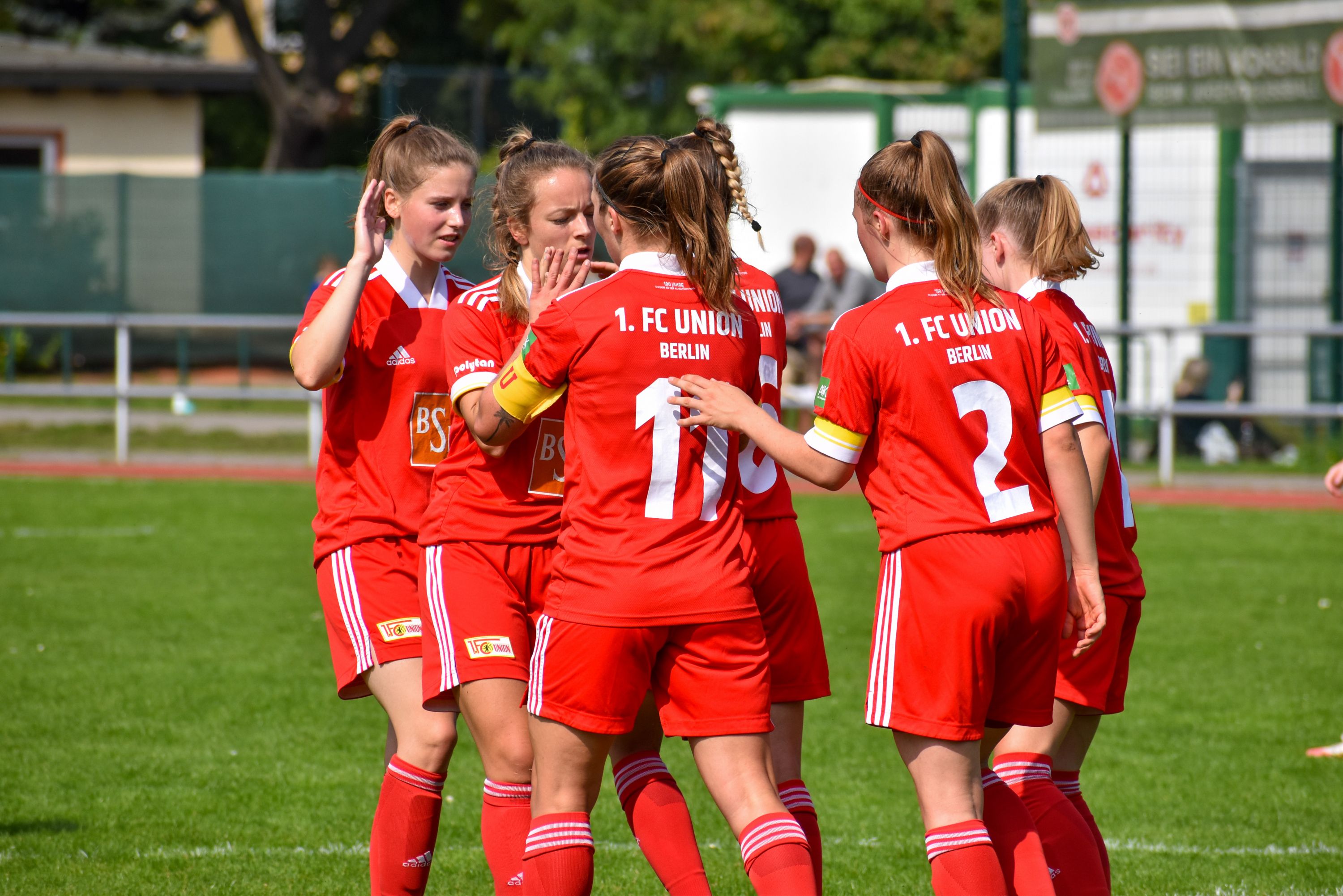 Fünf fußballspielende Frauen in roten Trikots feiern einen erfolgreichen Spielzug auf einem grünen Fußballfeld.